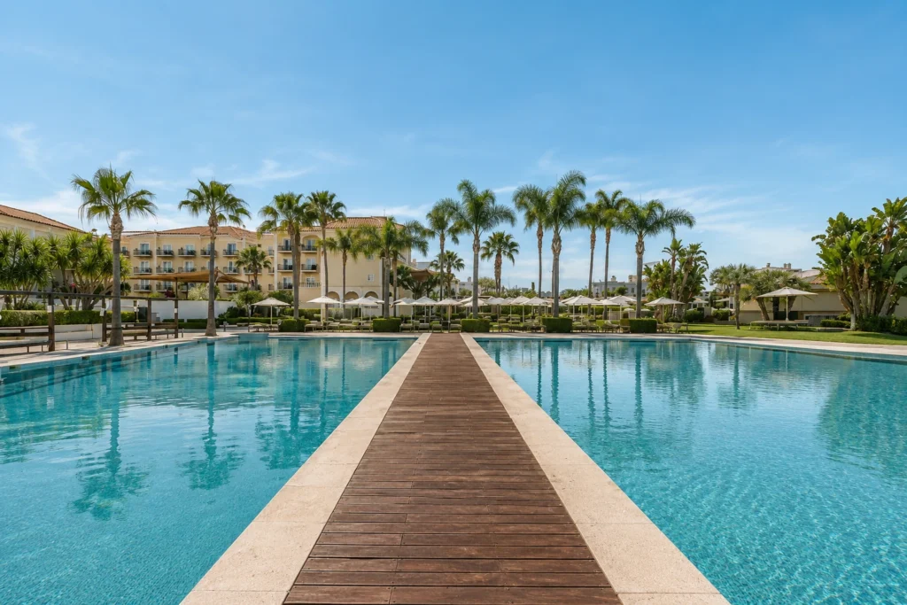 Daytime view of Domes Lake Algarve, Autograph Collection, showing a large outdoor pool with a wooden walkway, palm trees, sun loungers, and resort buildings under a clear blue sky.