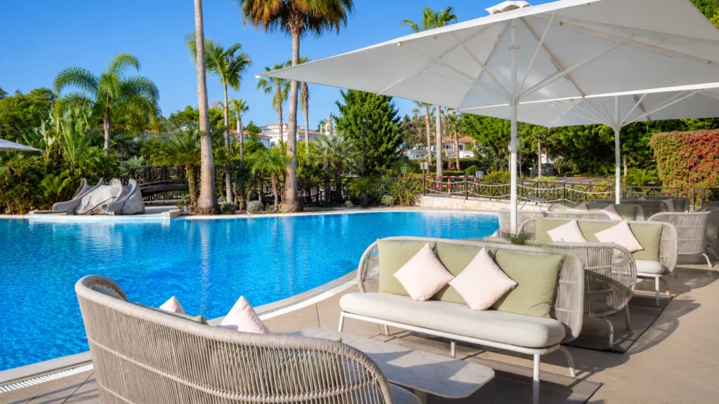 Daytime view of Martinhal Quinta do Lago Family Resort, showing a large outdoor swimming pool, cushioned seating under white parasols, tropical plants, palm trees, and a family-friendly resort setting.