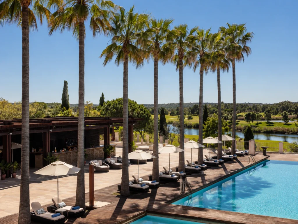 Daytime view of Victoria Golf Resort and Spa Managed by Accor, showing a large outdoor pool, palm trees, sun loungers, a stylish terrace bar, and golf-course scenery in the background.