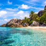 Seychelles beach with smooth granite boulders, palm trees, white sand and crystal-clear turquoise water under a bright blue sky.