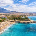 Tenerife coastal resort town with a sandy beach, turquoise sea, palm trees and mountain backdrop under a bright sky.