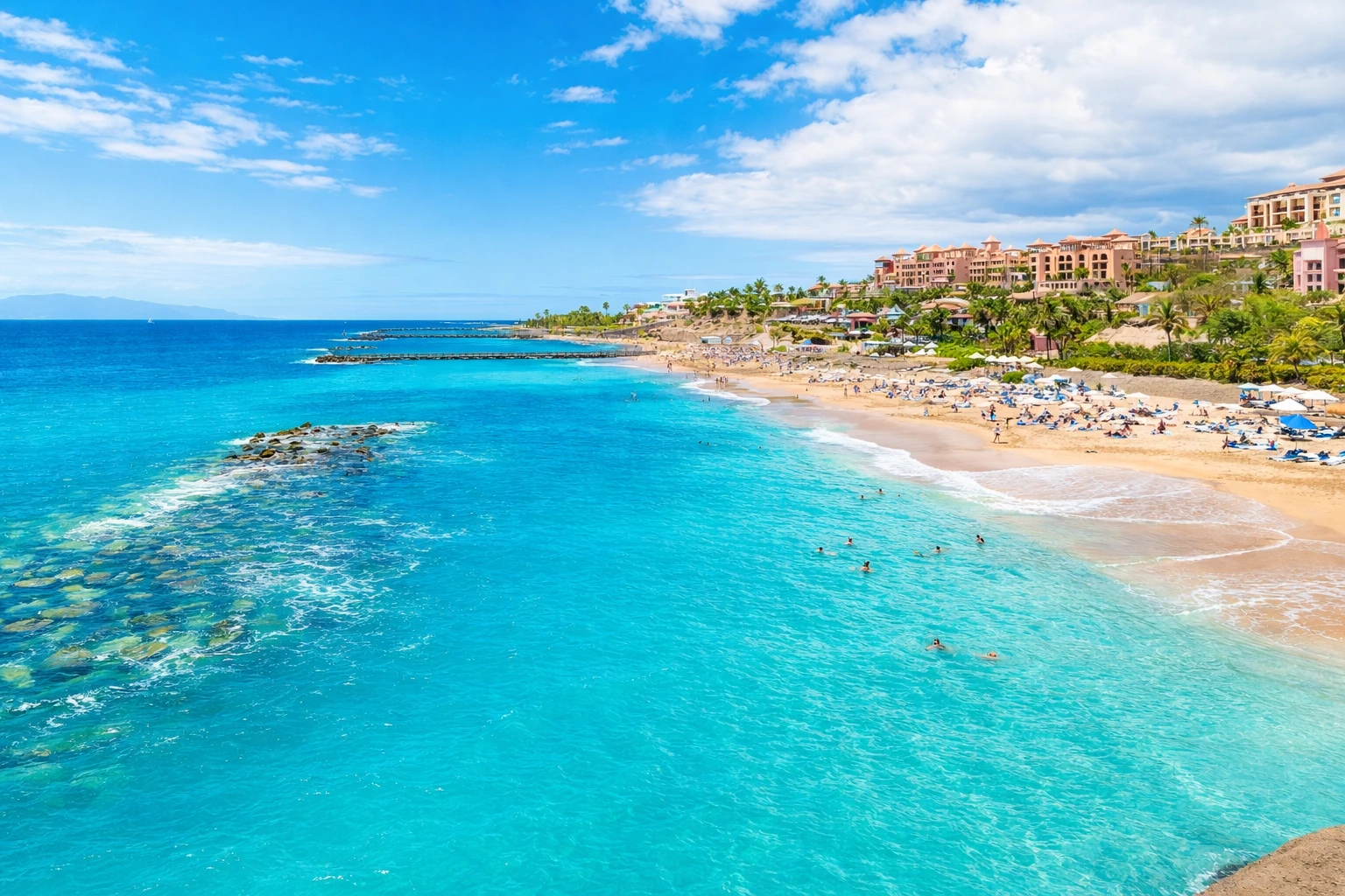 tenerife-luxury-family-resort-beach-escape Tenerife beach resort coastline with turquoise sea, sandy beach, luxury hotels and palm-lined waterfront under a bright blue sky.